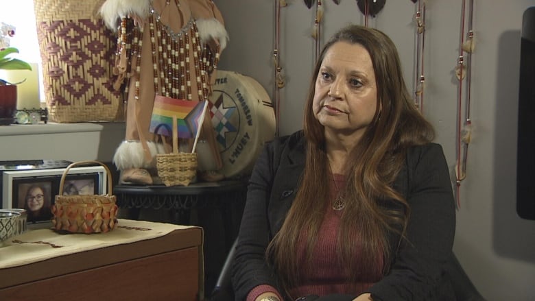 A woman with long brown hair sits in a room filled with Mi'kmaw baskets and ceremonial items. 