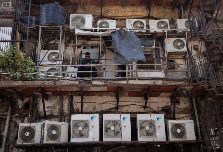 Several air conditioners line three levels of an apartment building and a man sits on a makeshift balcony.