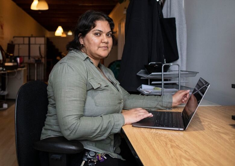 Meenakshi Mannoe looks at the camera while she is sitting down with a laptop. She is wearing an olive-coloured full-sleeved shirt.