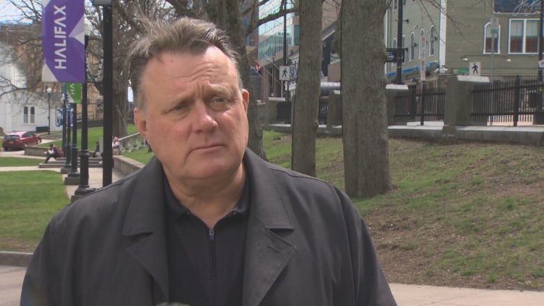 A white man in a black collared jacket and black shirt stands outside in a public square with trees behind him 