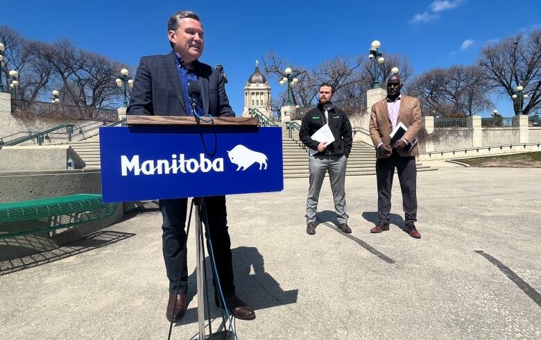 A man in a suit stands outside at a podium, with the Manitoba Legislature Building in the background. Two other men stand behind the man at the podium.