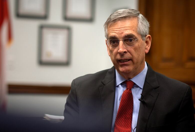 A man with grey hair wearing a suit speaks in a wood paneled room. 