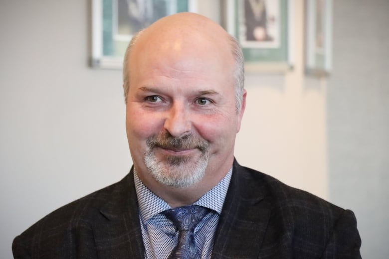 A man in a dark suit jacket with a blue shirt and dark blue tie with a short salt-and-pepper goatee smiles.