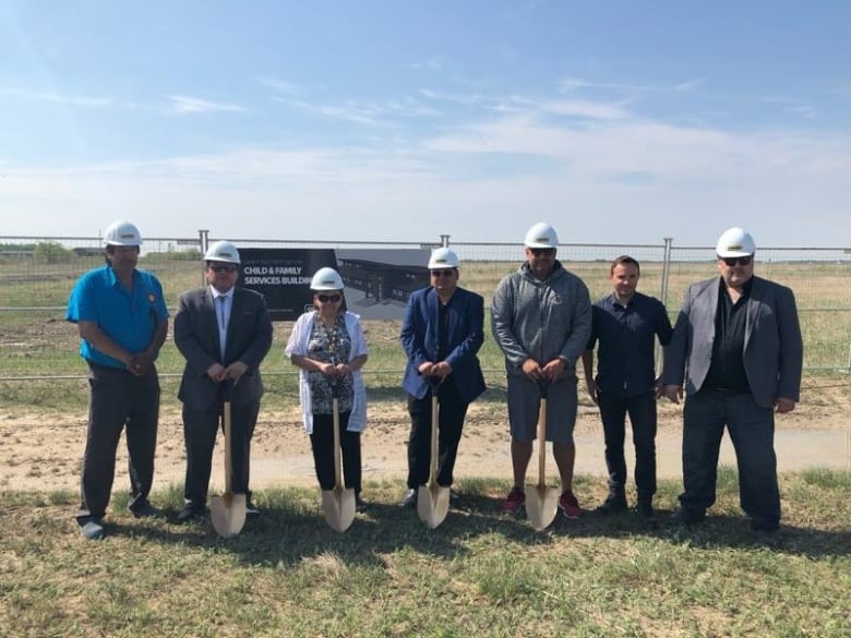 Seven people, six of them wearing white hard hats and four with shovels, stand in front of a fence in a field. A sign on the fence has an image of a building and says 