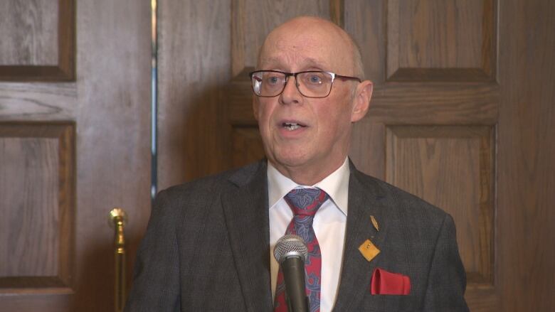 A man wearing glasses and a suit is speaking, facing the camera.