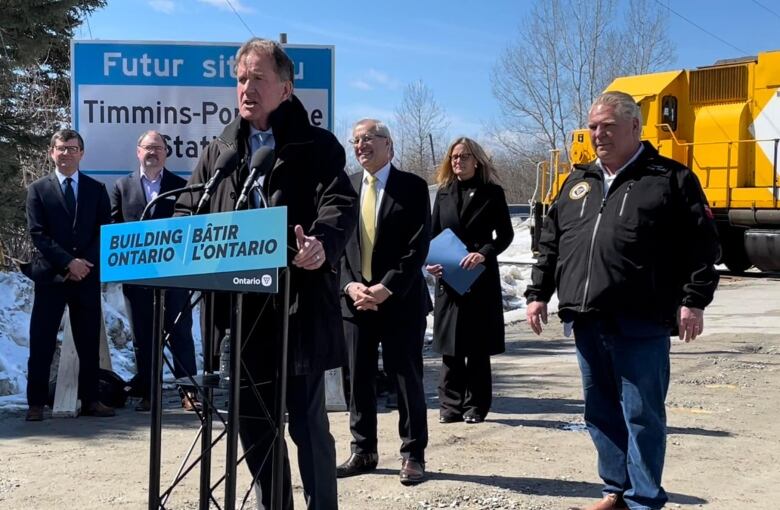 A politician stands at microphone and speaks, with others gathered behind him.