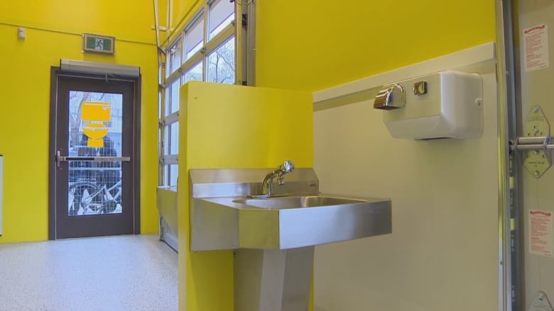 A stainless steel sink and a hand dryer are shown inside a washroom with bright yellow walls.