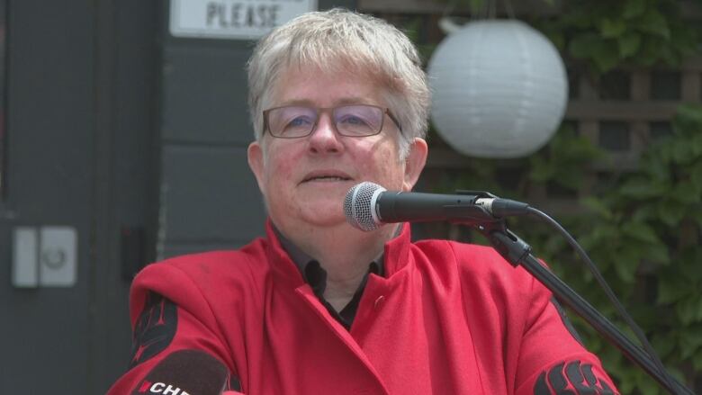 A woman with white/gray hair, wearing a red jacket and glasses, stands at a podium. 