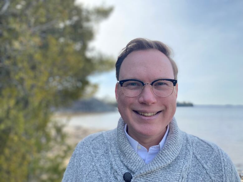 A man wearing a shawl-collared sweater and glasses smiles as he stands on the shore of a body of water.