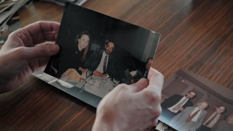 A pair of hands hold a photo of a bearded man and a woman at a dinner table.  