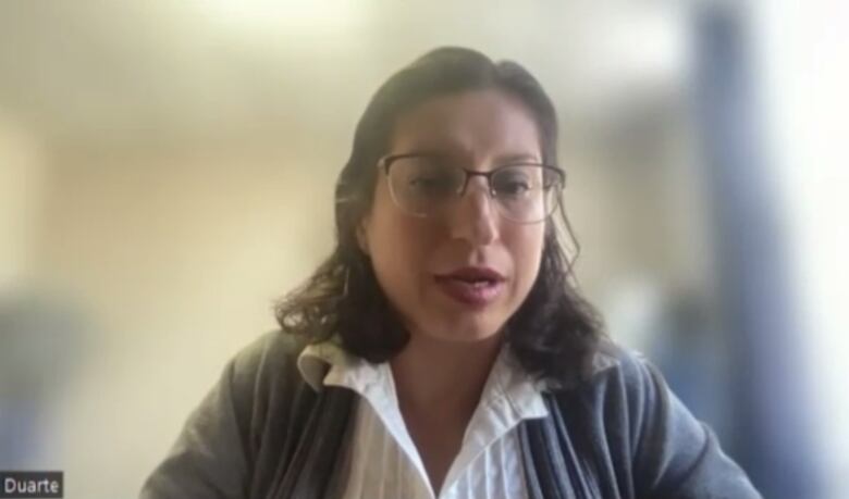 A woman with long dark curly hair and glasses speaks to a Zoom call.