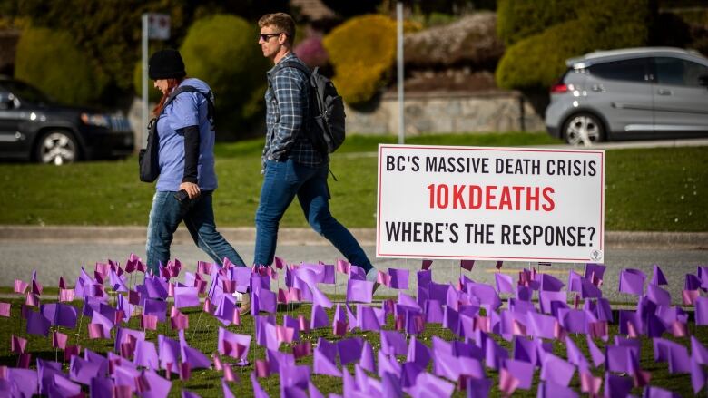 Two people walk by a sea of purple flags. A sign reads 'BC's Massive Death Crisis  10K Deaths  Where's the Response?'