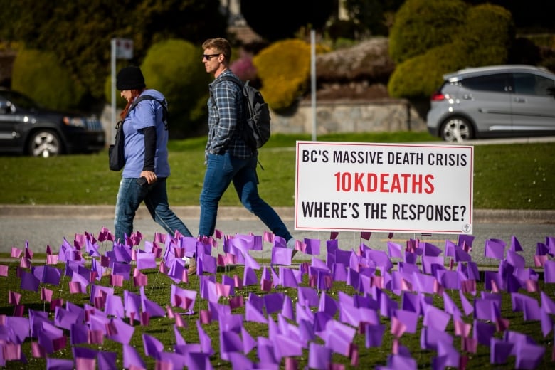 Two people walk by a sea of purple flags. A sign reads 'BC's Massive Death Crisis  10K Deaths  Where's the Response?'