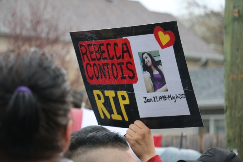 A person's hand is seen holding up a sign reading 'Rebecca Contois RIP.'