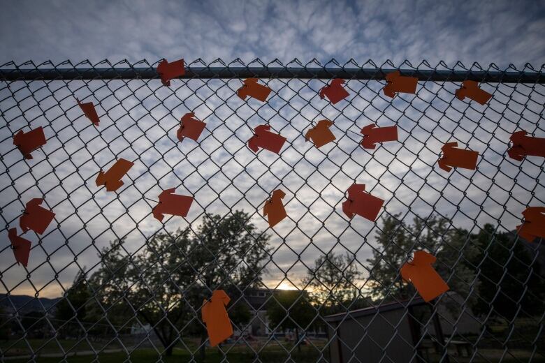 Several small orange shirts are clipped to a chain link fence, flapping in the wind as the sun sets behind it.