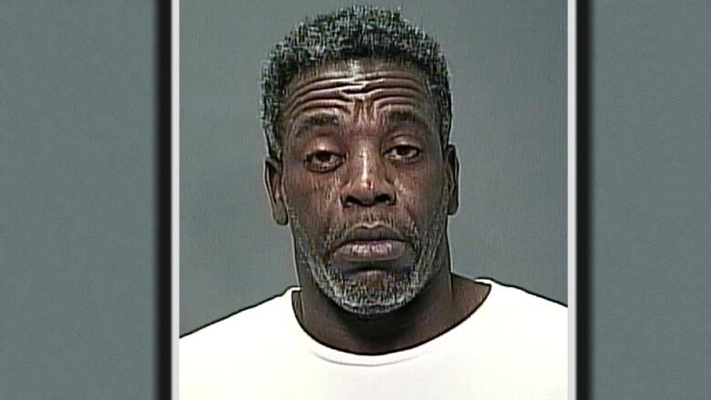 A man with a short black hair and black-and-grey stubble poses in a police mugshot.