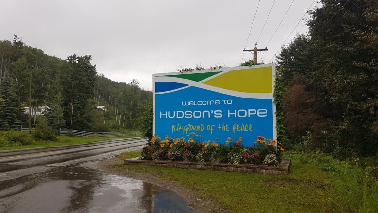 A green and blue sign by a roadway says, Welcome to Hudson's Hope.