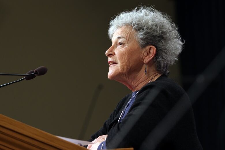 A woman with white, curly hair speaks into a mic at a podium. 