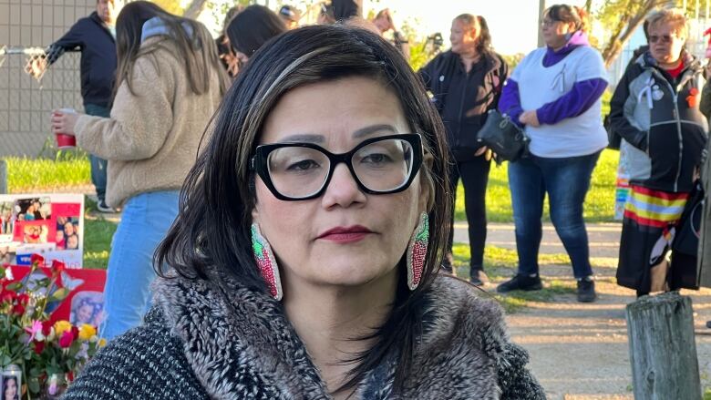 A woman with long dark hair wears bold, black-framed glasses and beaded earrings. She looks at the camera as other people stand in the background.