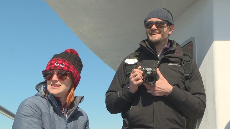 A man and a woman, both wearing tuques and sunglasses, are shown on a boat. The man holds a camera in his hands.