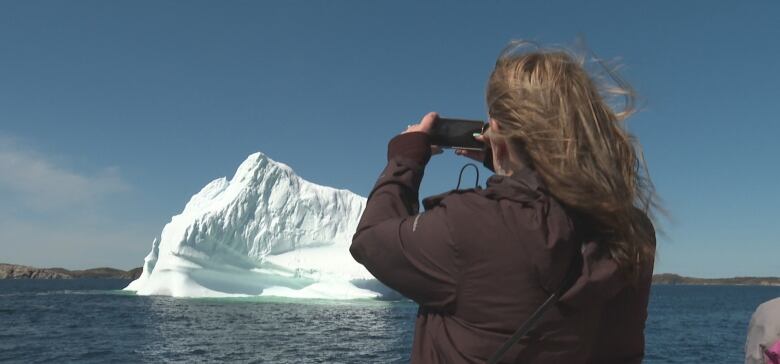 A woman wearing a black jacket holds up a cellphone to take a picture of a glacier.