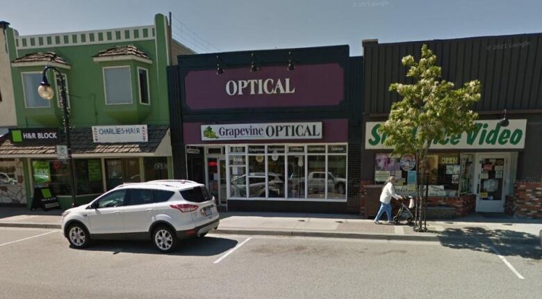 An optician's storefront on a street. The main sign, purple in colour, says 'Optical' in bold text. Underneath that, a smaller white sign says 'Grapevine OPTICAL' with a small clip art image of a bunch of grapes. It is next to a barbershop and a video shop.