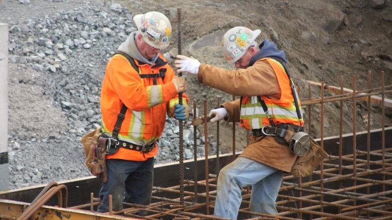 Construction workers in orange vests and white hard hats hold reinforcing bars in preparation for concrete to be poured into a wood frame.