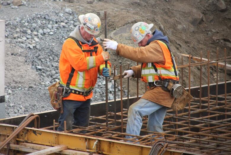 Construction workers in orange vests and white hard hats hold reinforcing bars in preparation for concrete to be poured into a wood frame.