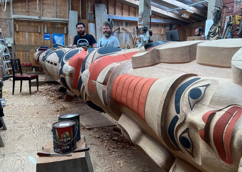 A large totem pole in progress lies on its side with two brothers standing near the head of the pole.