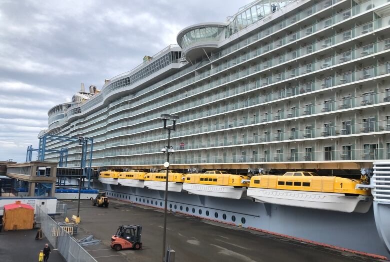 A very large cruise ship is docked. Lifeboats with yellow tops are on the lower side of the plaza.