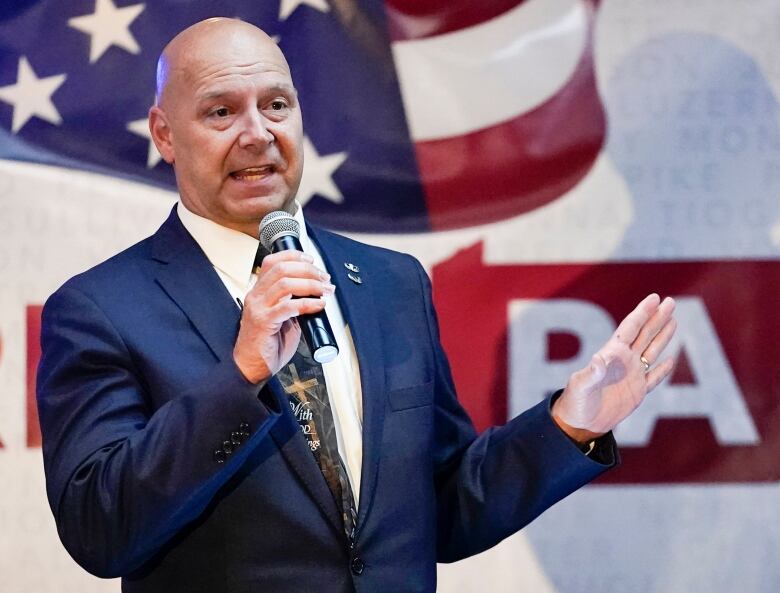 A bald middle aged man in a blue suit speaks into a microphone with the outline of Pennsylvania behind him.