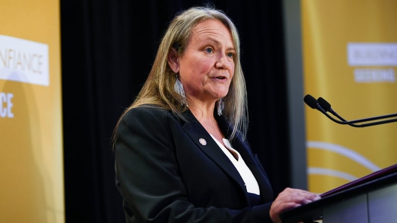 A woman with grey hair wearing a suit and beaded earrings speaks at a podium