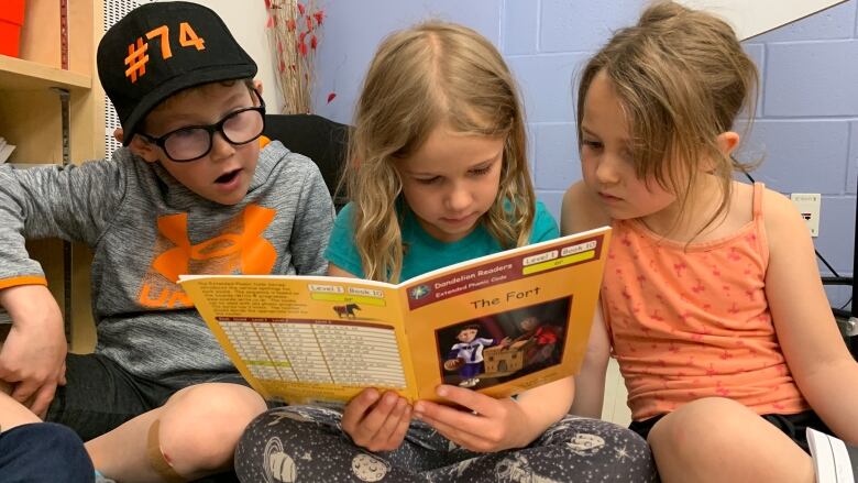 Three kids, each around six years old, sit cross-legged and side-by-side on the floor, looking at a picture book that the kid in the middle is holding open.