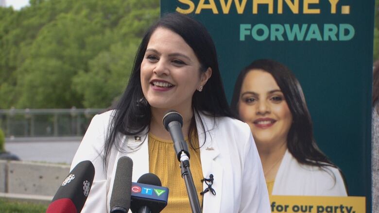 A woman with long black hair speaks into several microphones. She is standing in front of a giant photo of herself.