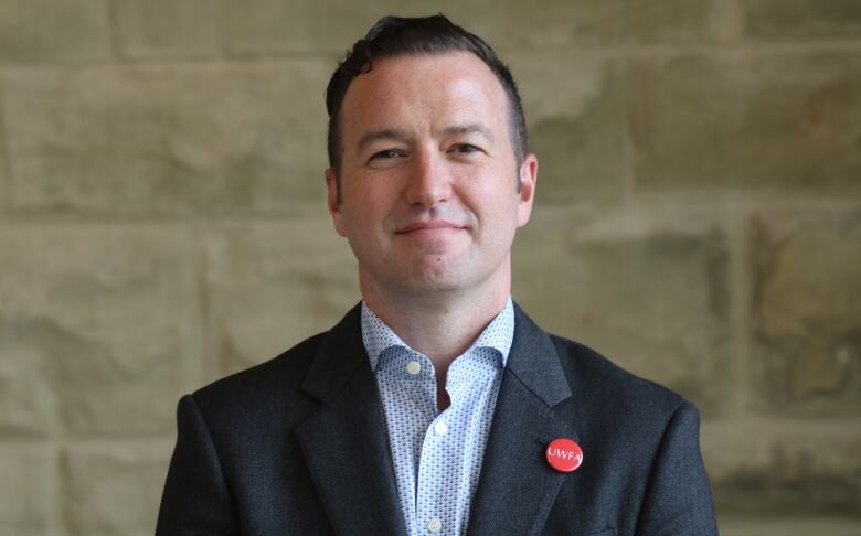 A man wearing a dark suit jacket with a red pin on the lapel reading 