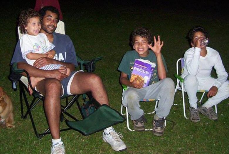 A dad and his three pre-teen kids sit on lawn chairs and smile at the camera.