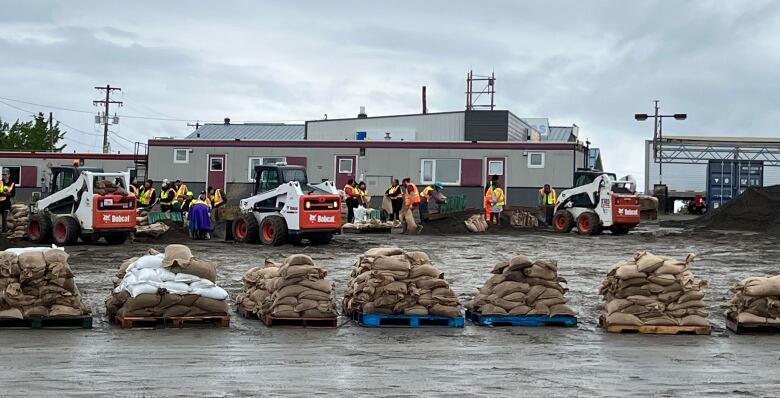 Sandbags are piled on pallets in a parking lot, with people working in the background.