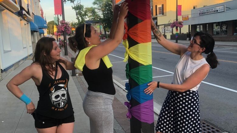 Three women tie rainbow-coloured strips of fabric to a light post.