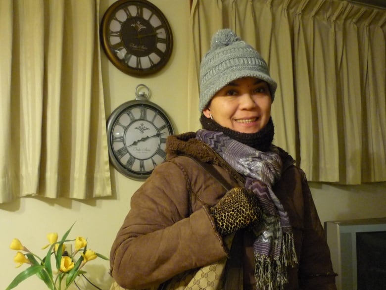 A Filipino woman in winter clothing stands with two clocks behind her.