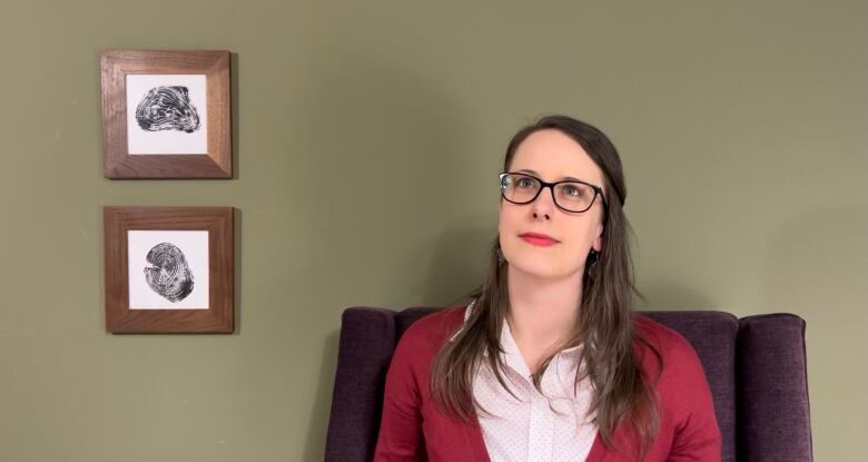 A white woman in her mid-30s with long, straight, dark brown hair, black plastic frame glasses, red lipstick, and a red cardigan over a pink button-up shirt, looks above and to the side of the camera with a small smile. Shes sitting in a purple arm chair in front of a light green wall that has two wood grain prints on it.