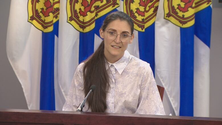 A woman in a light coloured print blouse with long hair and galsses sits in front of a number of blue-and-white Nova Scotia flags.