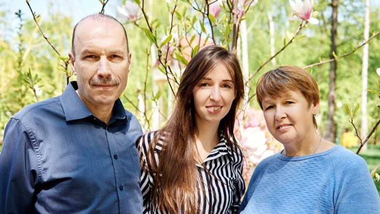 A smiling young woman is flanked by her parents, an older man and woman, posing for a photo in front of a flowering tree. 