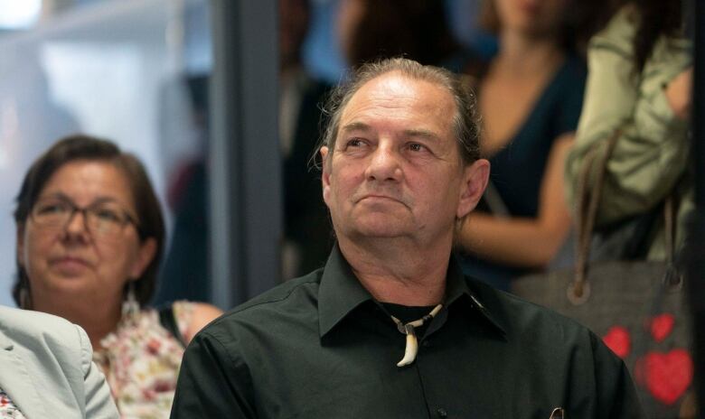 A man dressed in black and wearing a necklace with a tooth pendant listens.