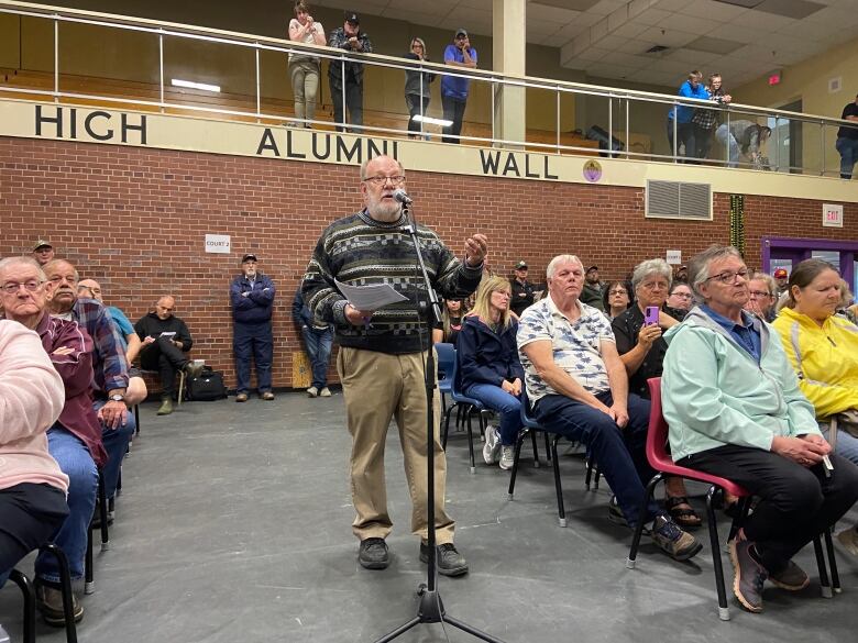 A many stands at a microphone at an outside event in front of people in people in chairs.