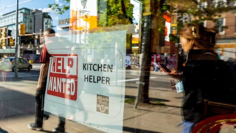 People walk past a help wanted sign adhered to a storefront window.