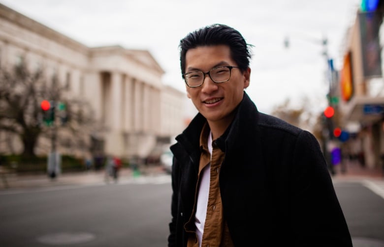 A man in a black jacket stands in front of a blurred out street.