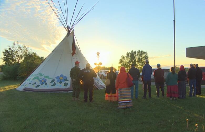 A crowd stands near a teepee as the sun rises.