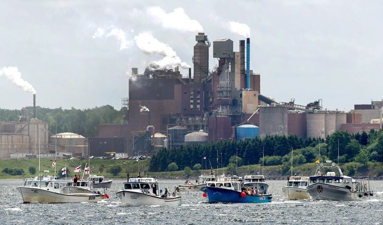 Boats on the water with a large factory on the shore in the background.