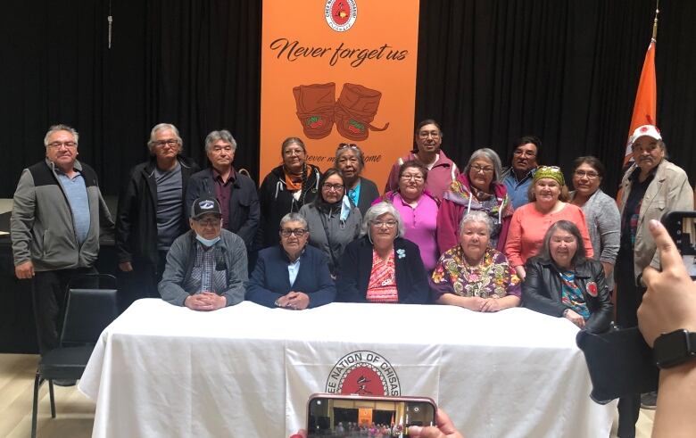 A group of men and women sit facing the camera. Behind them an orange banner reads: Never forget us.