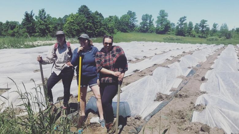 Three women hold shovels in front of some crops covered by landscaping fabric.